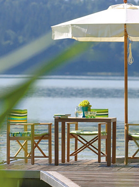 Wetterfeste Regiestühle Cabin aus Teakholz mit dem Stoff Spring als Sitzgruppe um einen Tisch und mit Sonnenschirm auf einem Steg am See.