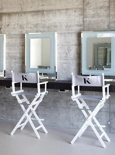 Zwei hohe Regiestühle Steven mit weißem Holzgestell und hellgrauem Stoff, inklusive Logodruck, stehen in einem Make-Up- und Beauty-Studio mit Spiegeln.