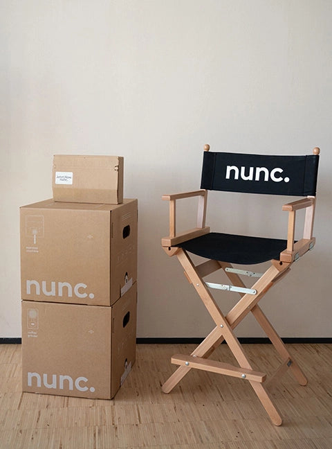 Hoher Regiestuhl Sydney mit einem Holzgestell in natur und schwarzem Stoff inklusive Logo-Druck steht neben gestapelten Verpackungskartons.