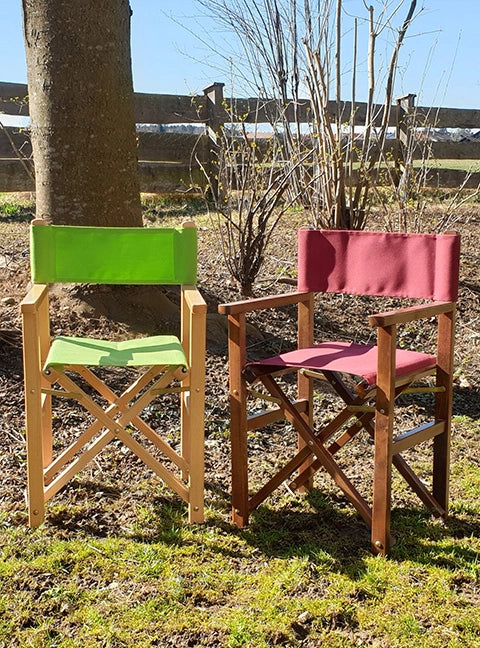 Zwei Kinderregiestühle Nanni in den Ausführungen natur Holz mit hellgrünem Stoff und braunes Holz mit Fuchsia Stoff stehen im Garten.