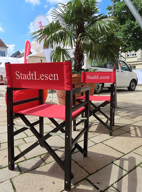 Zwei Regiestühle Oscar in schwarz lackiertem Holz mit rivarotem Stoff und Logo-Druck stehen auf einem öffentlichen Platz für ein Event.