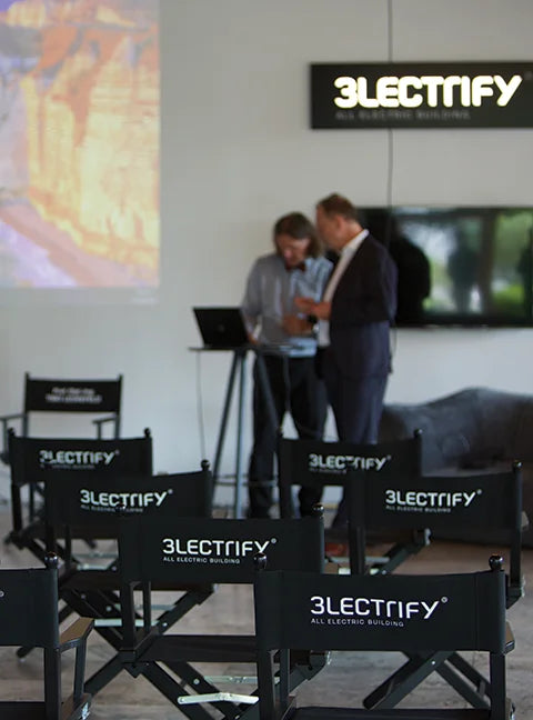 Mehrere schwarze Regiestühle Alfred mit 3LECTRIFY-Logo in Reihenbestuhlung bei einem Event mit Präsentation und den Speakern im Hintergrund.