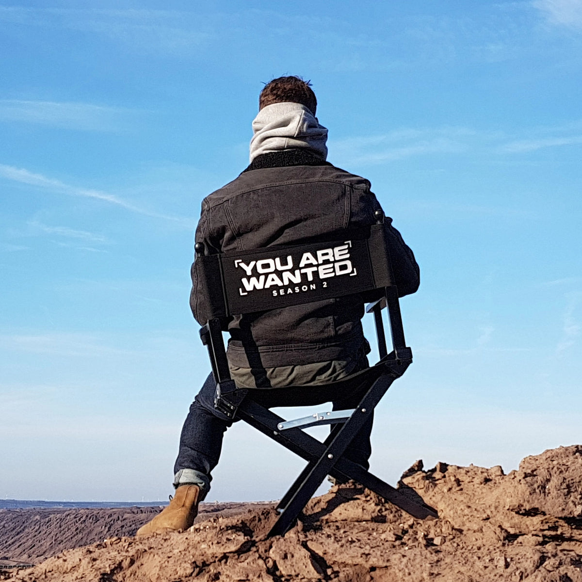Schwarzer Regiestuhl Alfred mit Logo-Aufdruck ‚You Are Wanted‘ bei Outdoor-Dreh in Film- oder TV-Produktion – ideal für Set-Design, Regie und Crew-Ausstattung