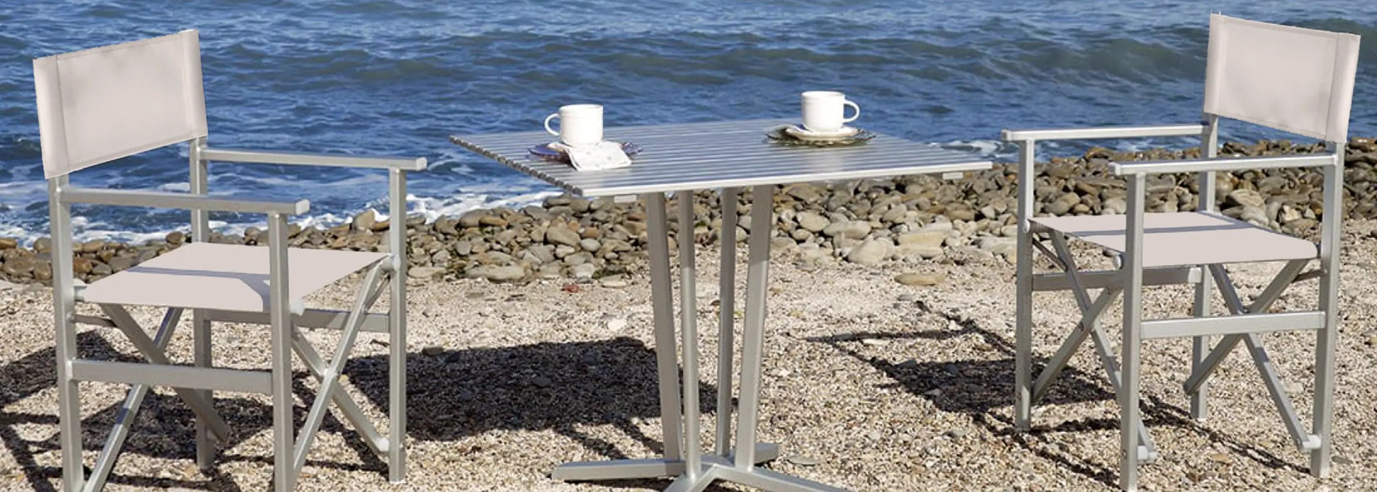 Zwei Regiestühle Stanley aus Aluminium mit hellgrauem Stoff auf einem Strand mit Steinen und Blick aufs Meer.