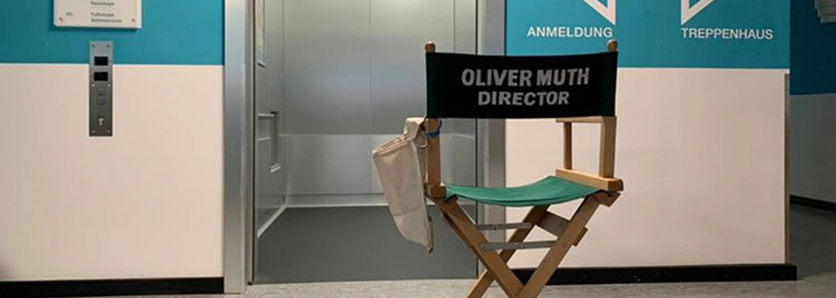 Regiestuhl mit dunkelgrünem Bezug und Aufschrift „Oliver Muth – Director“ vor einem Aufzug in einer Filmklinik-Location.