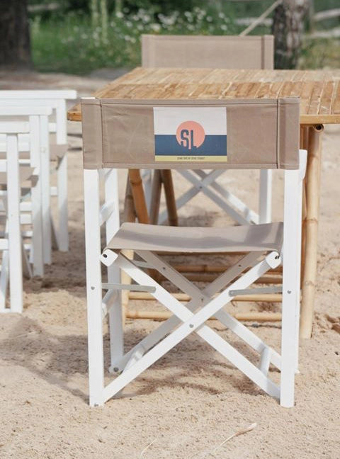 Regiestuhl Oscar mit weißem Gestell und grau-beigem Bezug samt individuellem Logo-Druck in einer Strandbar im Einsatz.