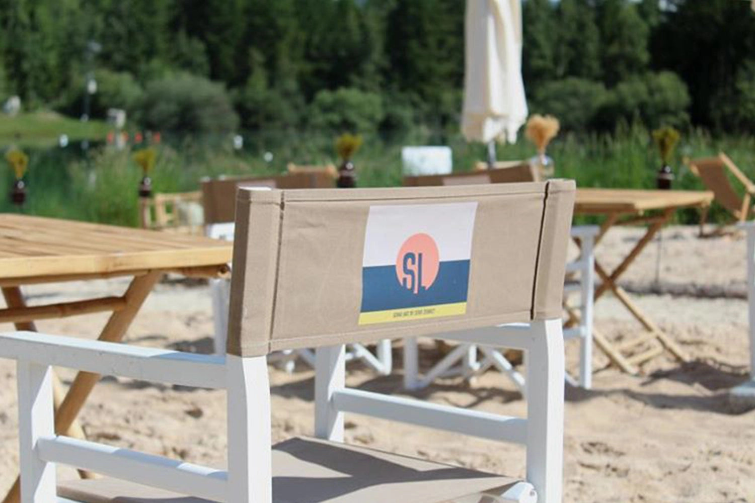 Regiestuhl Oscar mit weißem Gestell und grau-beigem Stoff inklusive Logo-Druck platziert im Sandstrand-Ambiente einer Outdoor-Location.