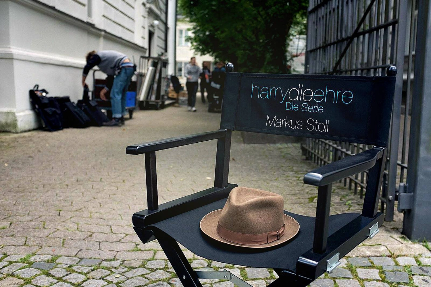 Schwarzer Regiestuhl Steven mit Logodruck ‚harrydieehre – Die Serie‘ und braunem Hut auf dem Sitz – im Einsatz bei einem TV-Dreh vor urbaner Kulisse.