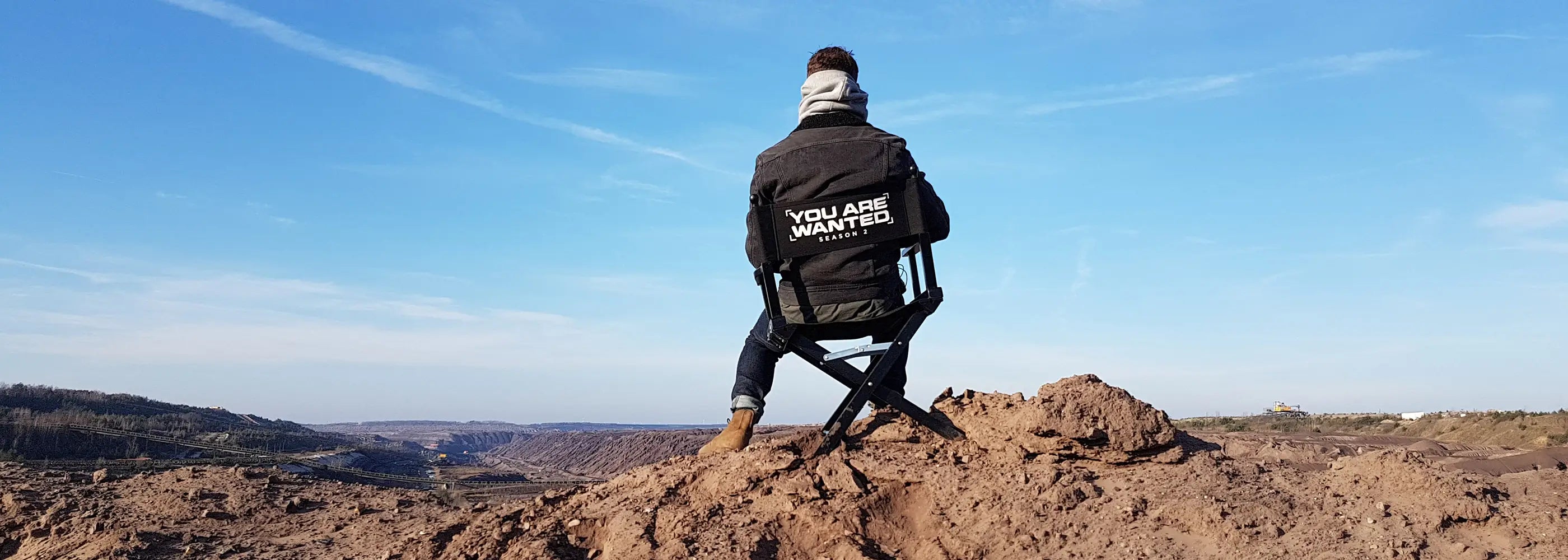 Schauspieler sitzt auf Regiestuhl mit Aufschrift „You Are Wanted – Season 2“ in beeindruckender Landschaftskulisse.
