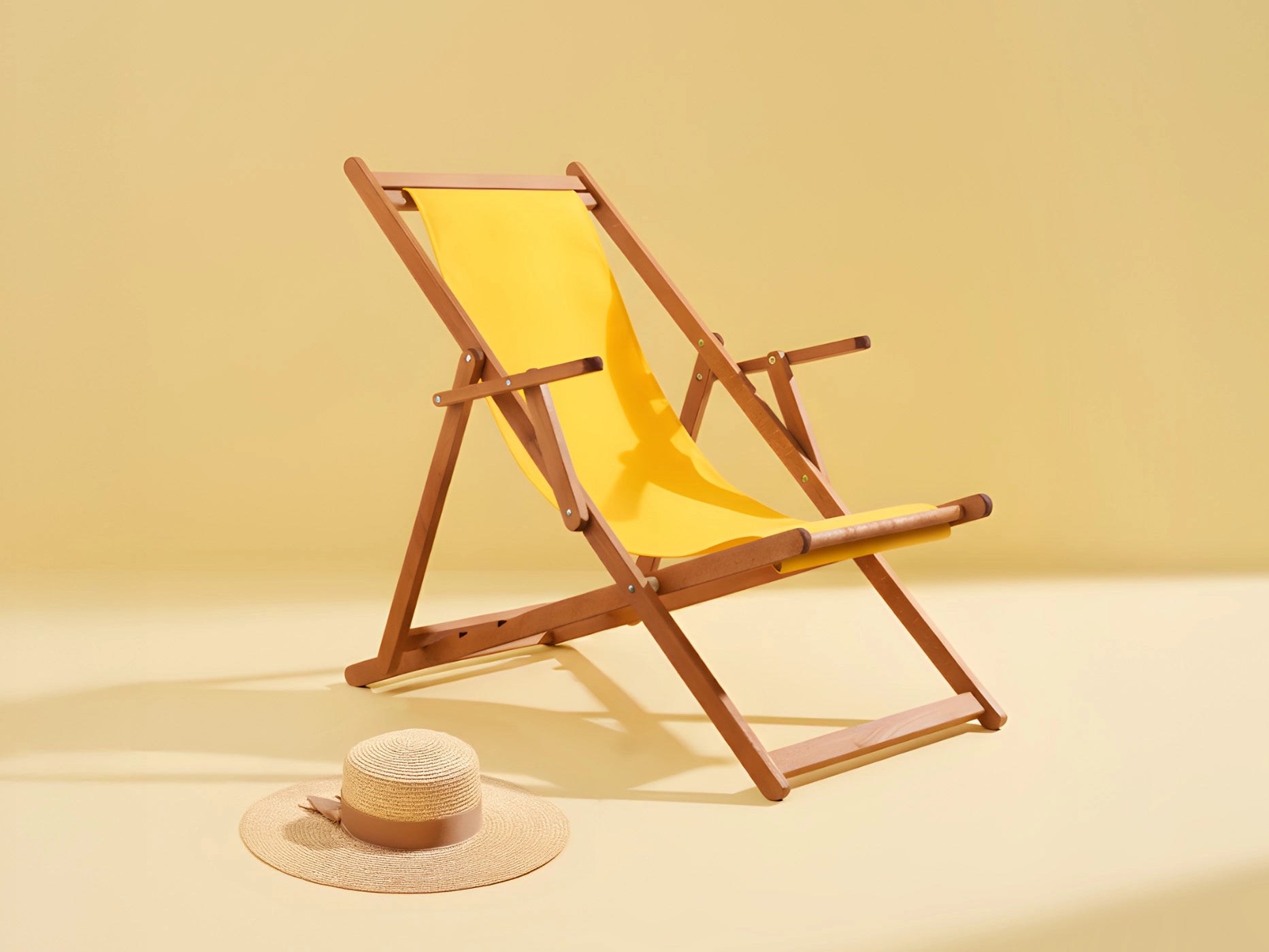 Gelber Liegestuhl aus Holz mit Sonnenhut daneben vor monochrom gelbem Hintergrund