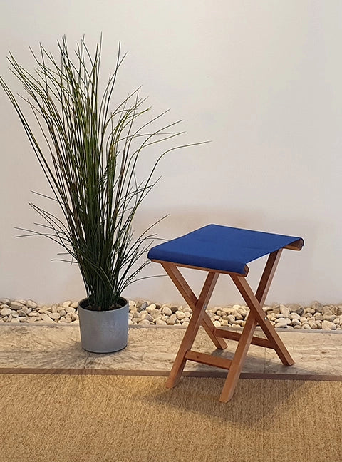 Beistellhocker Wim aus Holz in brandy mit dunkelblauem Bezug, platziert neben einer hohen Pflanze im modernen Wohnambiente