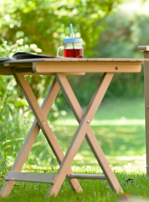 Beistelltisch Roland in naturbelassenem Holz, darauf ein Erfrischungsgetränk und Zeitschrift, steht in einem grünen Garten.