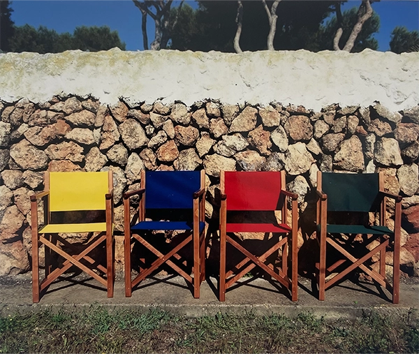 Vier Regiestühle Pedro in brandy mit rotem, gelben, blauen und dunkelgrünem Stoff vor einer menorquinischen Steinmauer – Symbol für die frühe Farbvielfalt und den mediterranen Stil der Marke.