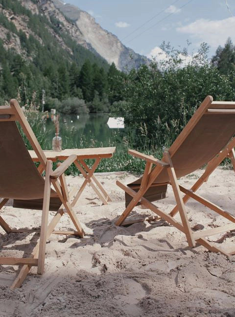 Liegestühle Charly mit natur Gestell und grau-beigem Bezug von hinten fotografiert an einem Strand am See, davor zwei Beistelltische Roland in natur.