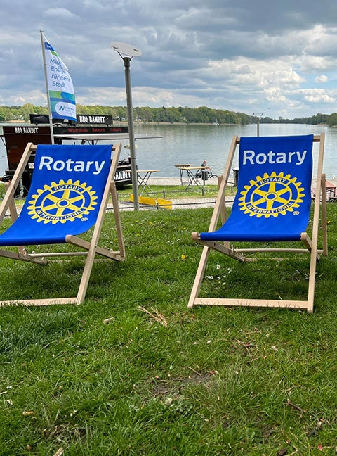 Zwei personalisierte Liegestühle Oliver aus naturbelassenem Holz und mit Logo-Druck stehen auf einer Wiese an einem See.