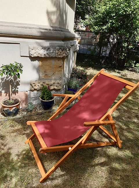 Liegestuhl Charly aus Holz in brandy geölt, mit Armlehnen und einem Stoff in Bordeaux im Garten stehend.