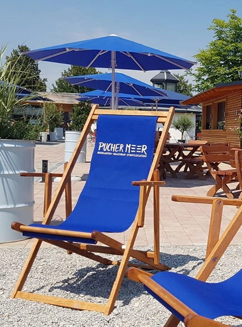 Liegestuhl Charly aus Holz in brandy geölt, mit Armlehnen und einem Stoff in dunkelblau inklusive Logo-Druck für den Einsatz im Außenbereichs eines Gastronomiebetriebs.