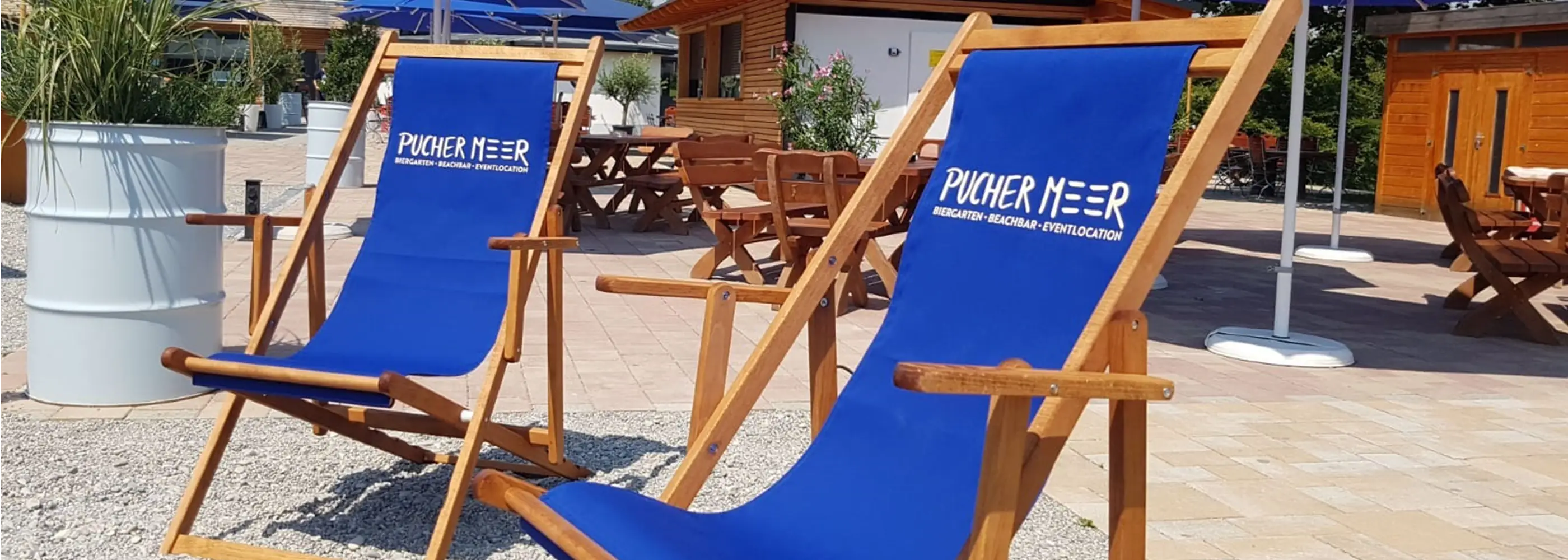 Zwei Liegestühle Charly aus Holz mit dunkelblauem Stoff und Logo-Bedruckung im Außenbereich einer Eventlocation mit Sonnenschirmen.