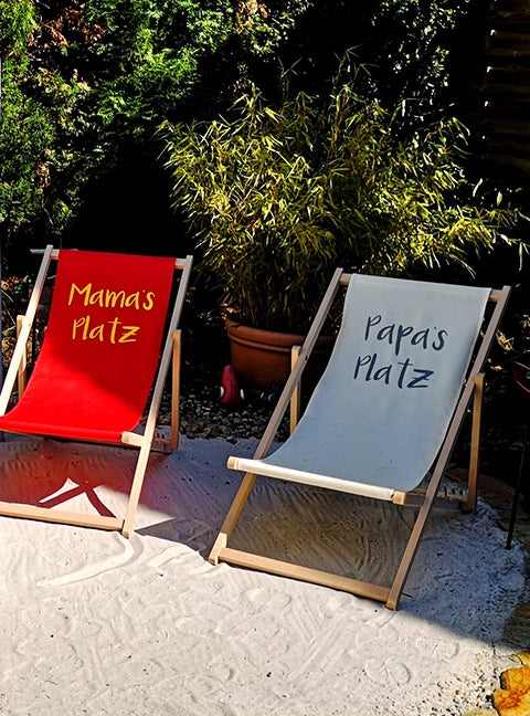 Zwei Liegestühle Oliver aus naturbelassenem Holz und mit personalisiertem Aufdruck sind im Garten aufgestellt.