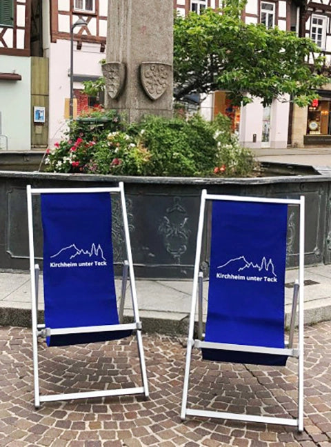 Aluminium Liegestuhl Quentin in silber mit dunkelblauem Stoff und Logo-Bedruckung auf einem öffentlichen Platz vor einem Brunnen.