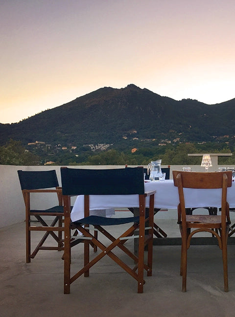 Regiestühle Woody in braun mit schwarzen Bezügen an einem gedeckten Esstisch auf einer Dachterrasse mit Blick auf mediterrane Berge bei Sonnenuntergang