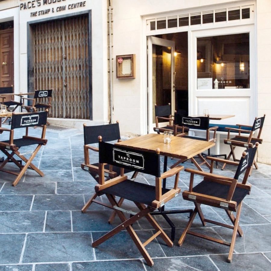 Regiestühle Alfred aus braunem Holz mit schwarzem Bezug und Logo-Druck, stilvoll arrangiert in einer Gasse vor einem Straßencafés – ideal für Restaurants, Bistros und Hotels.