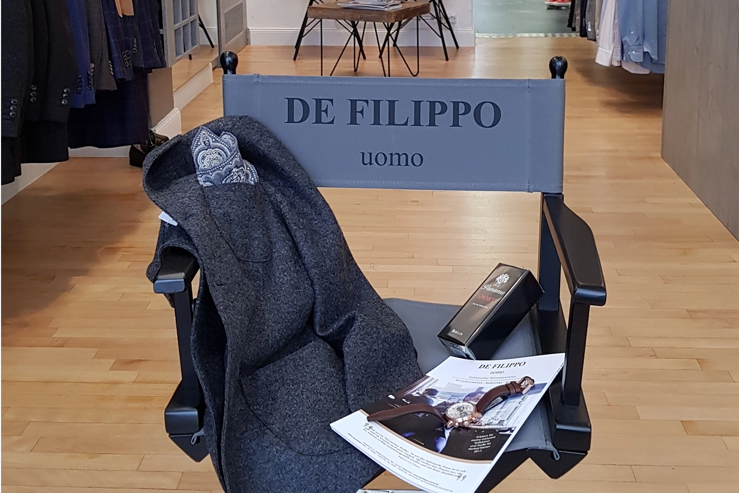 Regiestuhl Alfred mit schwarzem Holzgestell und anthrazitem Bezug mit Logo-Druck, arrangiert in einer Herrenboutique mit Mantel, Accessoires und Modezeitschriften.