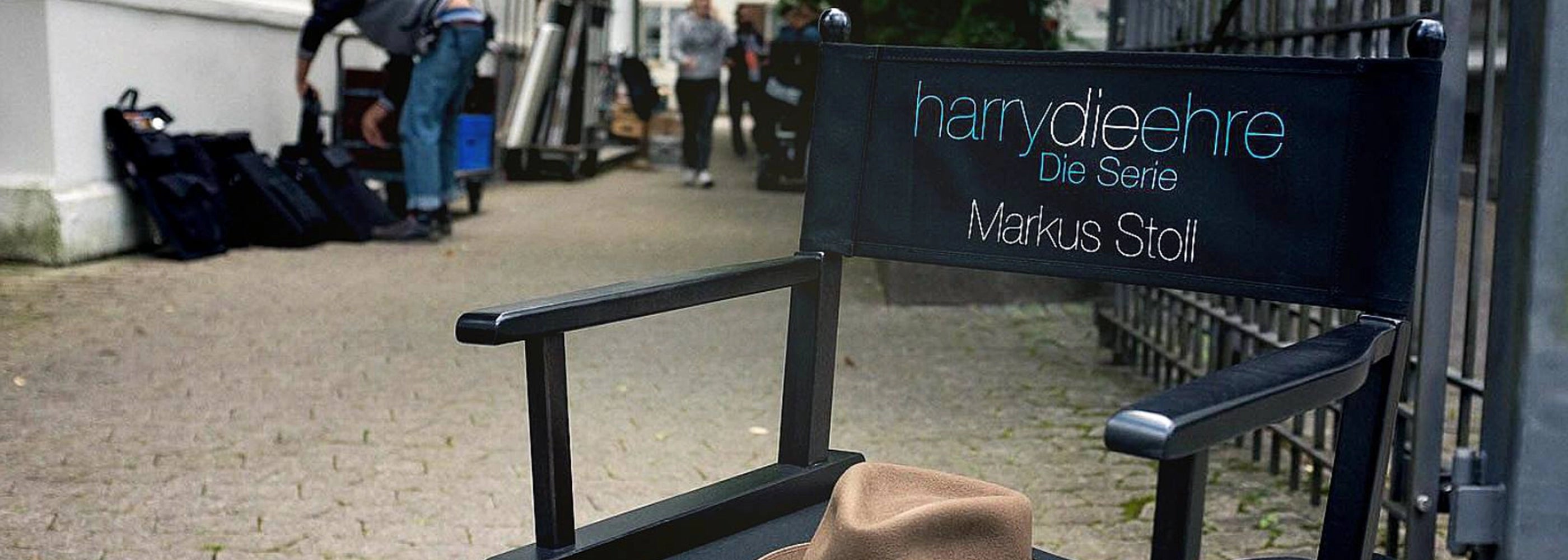 Regiestuhl mit Aufdruck „harrydieehre – Die Serie, Markus Stoll“ am Set, im Hintergrund das Filmteam.