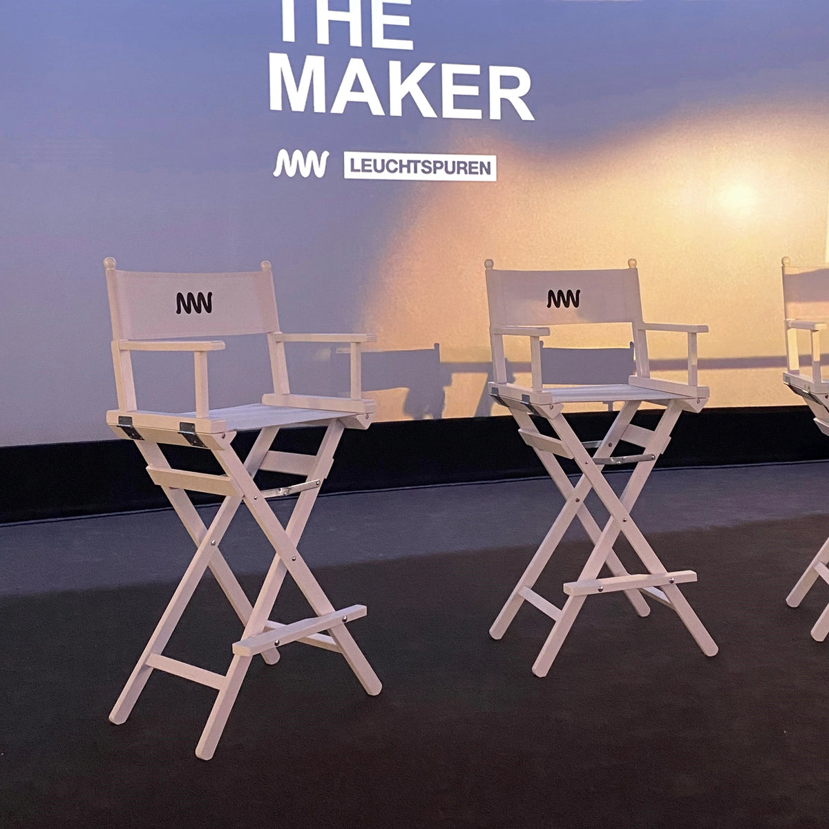 Hohe Regiestühle Steven mit weißem Holzgestell und Logo-Druck im Einsatz bei einer Podiumsdiskussion auf einem Business-Event mit Messecharakter – stilvolle Sitzlösung für Talks & Panels