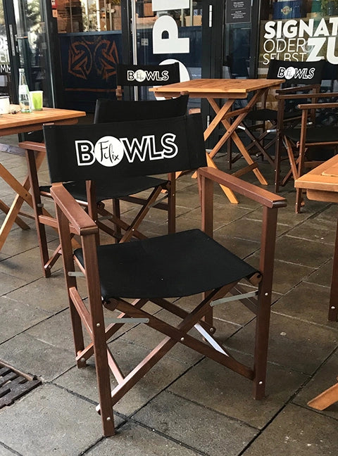 Regiestuhl Woody in braun mit schwarzem Bezug und Logodruck wird auf einer Restaurant-Terrasse eingesetzt.