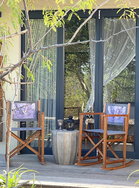 Zwei Schaukelstühle Helmut mit Holz in brandy und Stoff in Anthrazit mit einem Fotodruck stehen auf einer Veranda einer Lodge in Afrika.